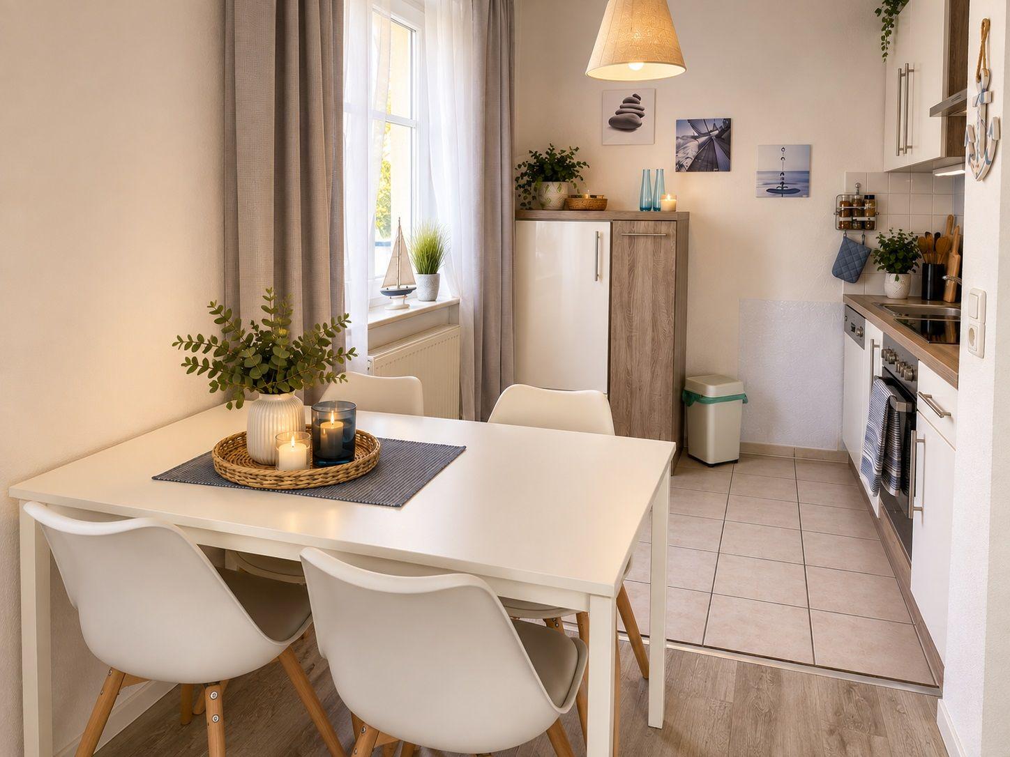 Essbereich mit weißem Tisch und vier Stühlen. Fenster mit Vorhängen und Küchenmöbel im Hintergrund.