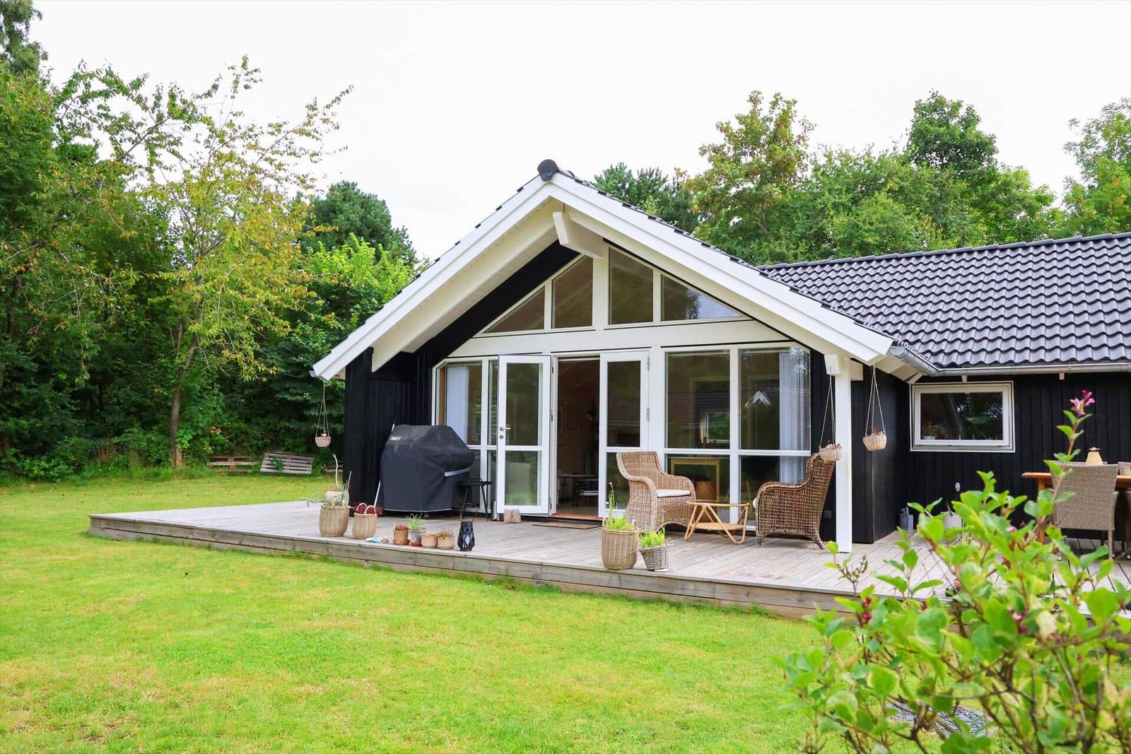Terrasse mit Rattanmöbeln, Holztisch und Pflanzenkörben. Hintergrund: Haus mit großen Fenstern.