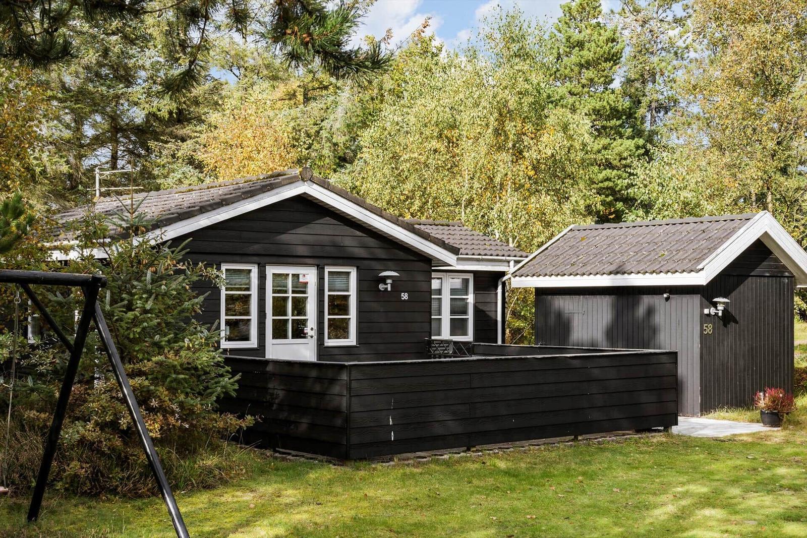 Black wooden house with white windows and paved yard. Surrounded by trees.