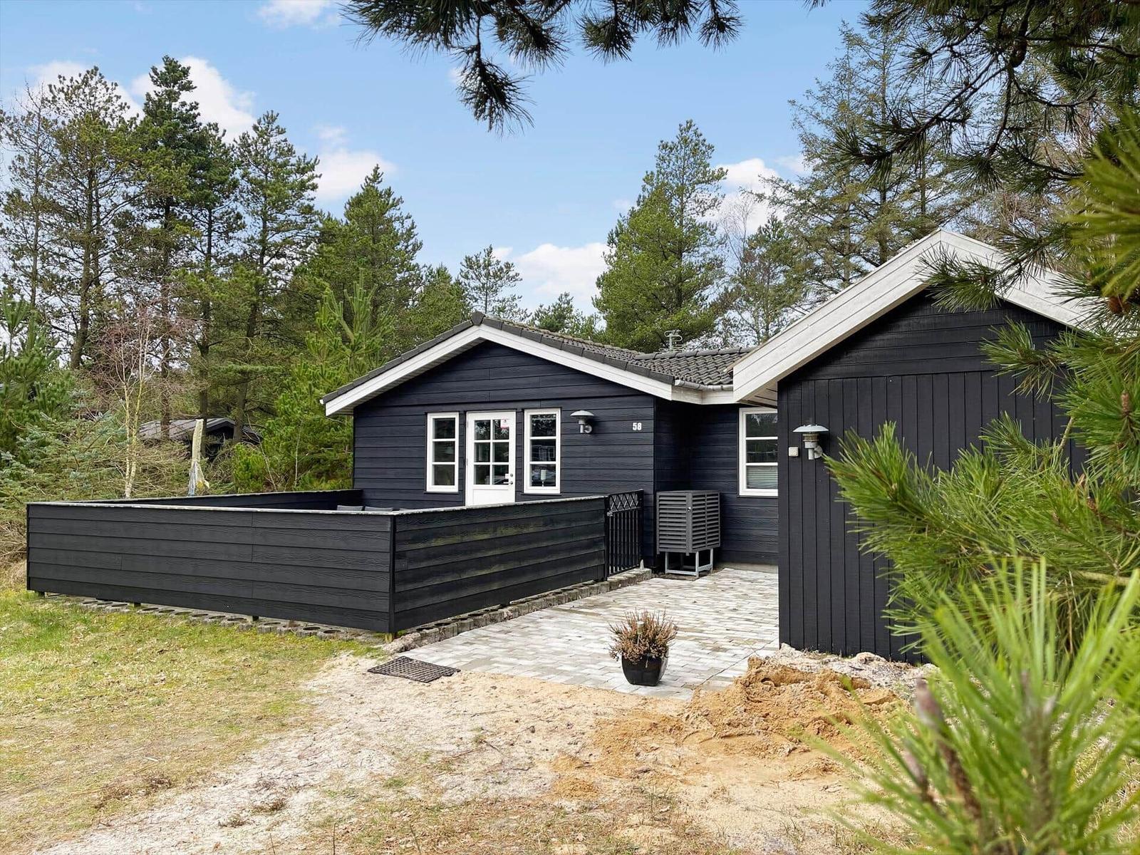 Black wooden cabin with terrace, table, and chairs. Surrounded by trees and grass.