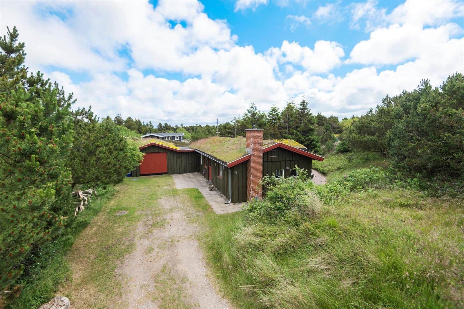 Holzhaus mit grüner Dachdeckung, roter Tür und Schornstein. Umgeben von Gras und Bäumen.