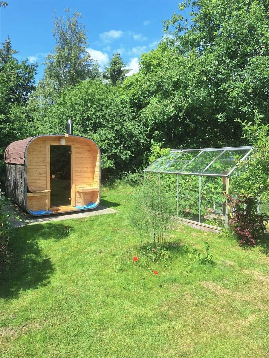 Holzsauna und Gewächshaus im grünen Garten mit Bäumen und Rasen.
