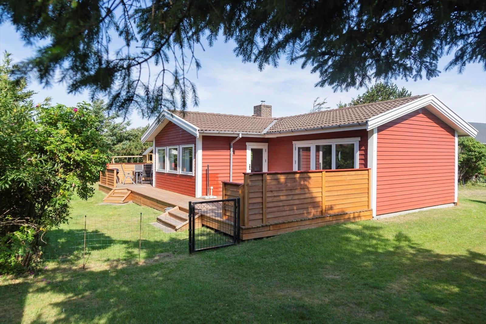 Rotes Holzhaus mit Terrasse und Garten. Holzgeländer und Schiebetüren sind sichtbar.