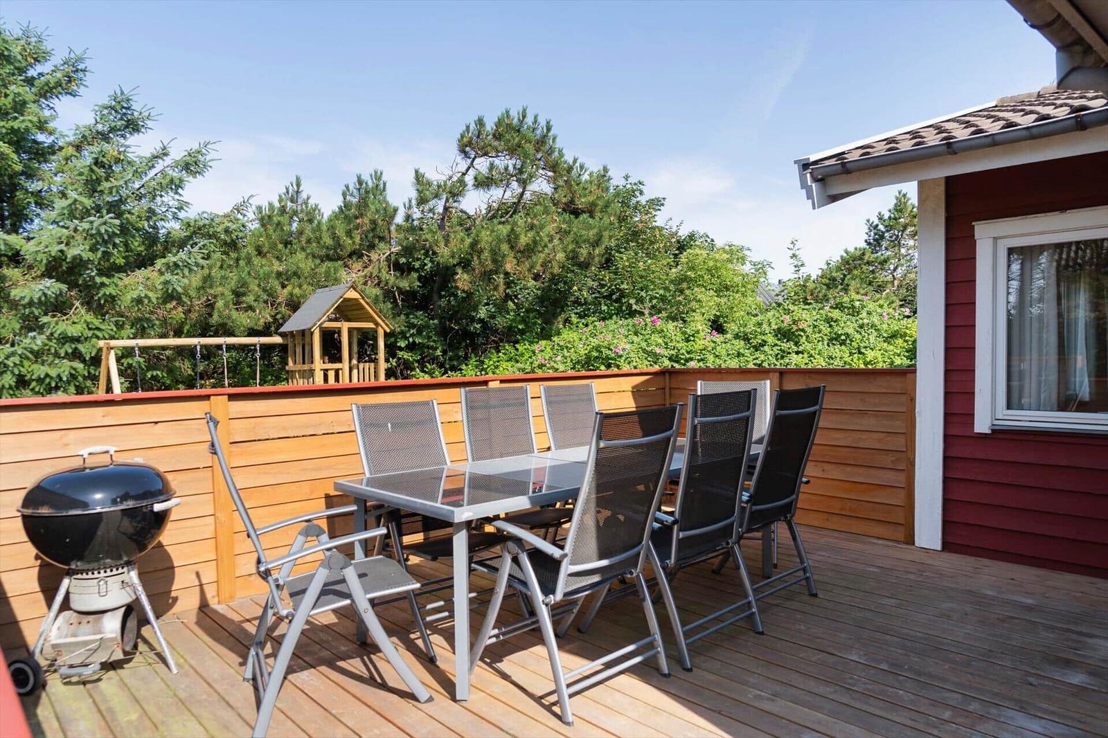 Terrasse mit Grill, Tisch und sechs Stühlen. Hintergrund: Holzzaun und grüne Bäume.