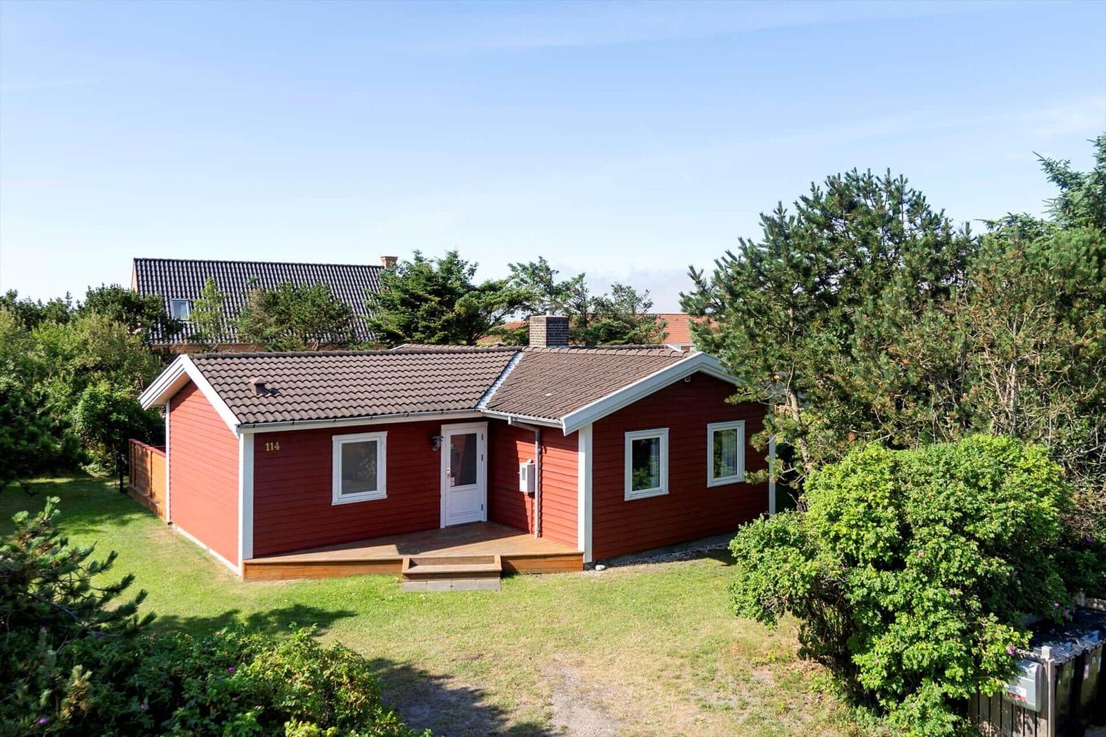 Rotes Haus mit weißen Fensterrahmen und Vorgarten. Hintergrund mit Bäumen und klarem Himmel.