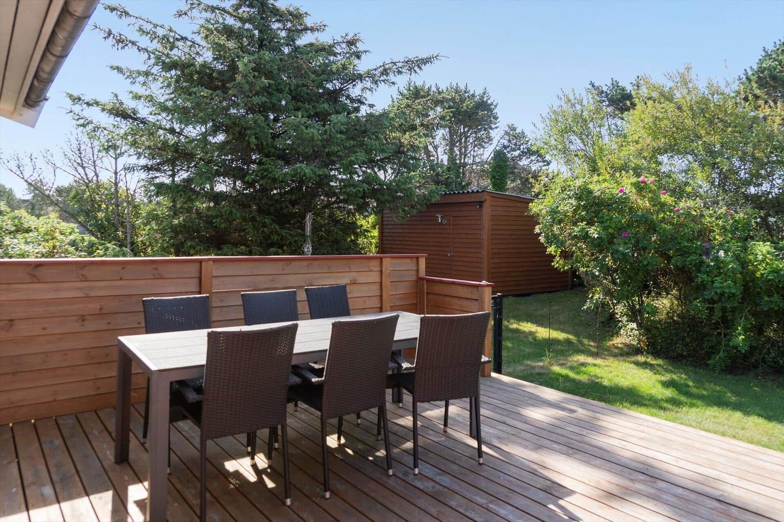 Terrasse mit Tisch und sechs Stühlen, umgeben von Grün und Holzgeländer.