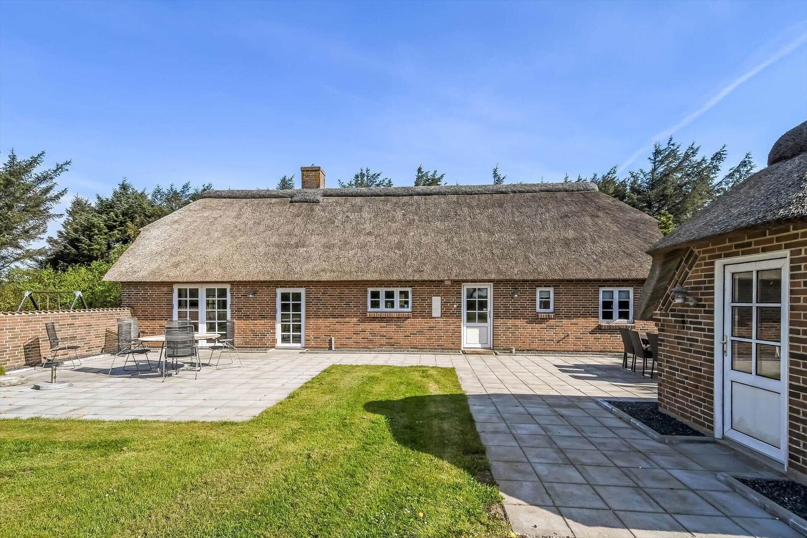 Dorfhaus mit Dach aus Stroh, umgeben von Grünflächen und Bäumen, mit Blick auf die Dünen und das Meer.