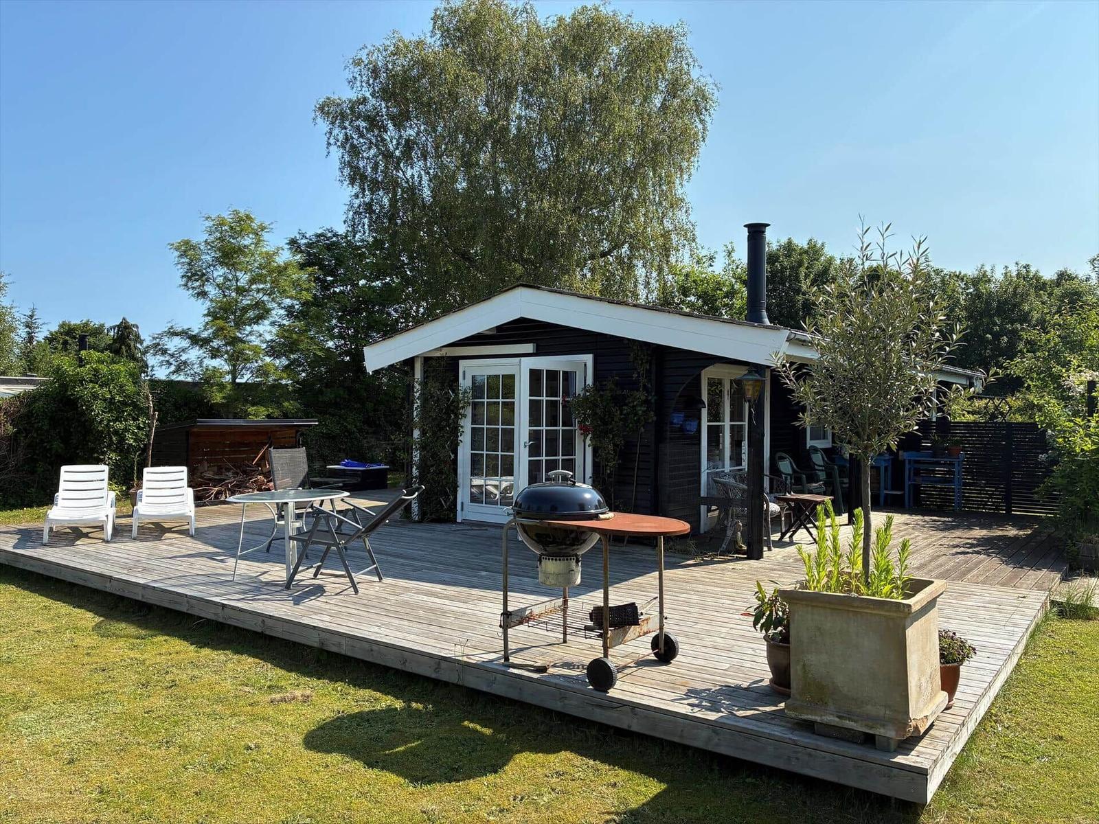 Schwarzes Holzhaus mit Garten, Bank und Blumen. Rauchrohr und Bäume sind sichtbar.