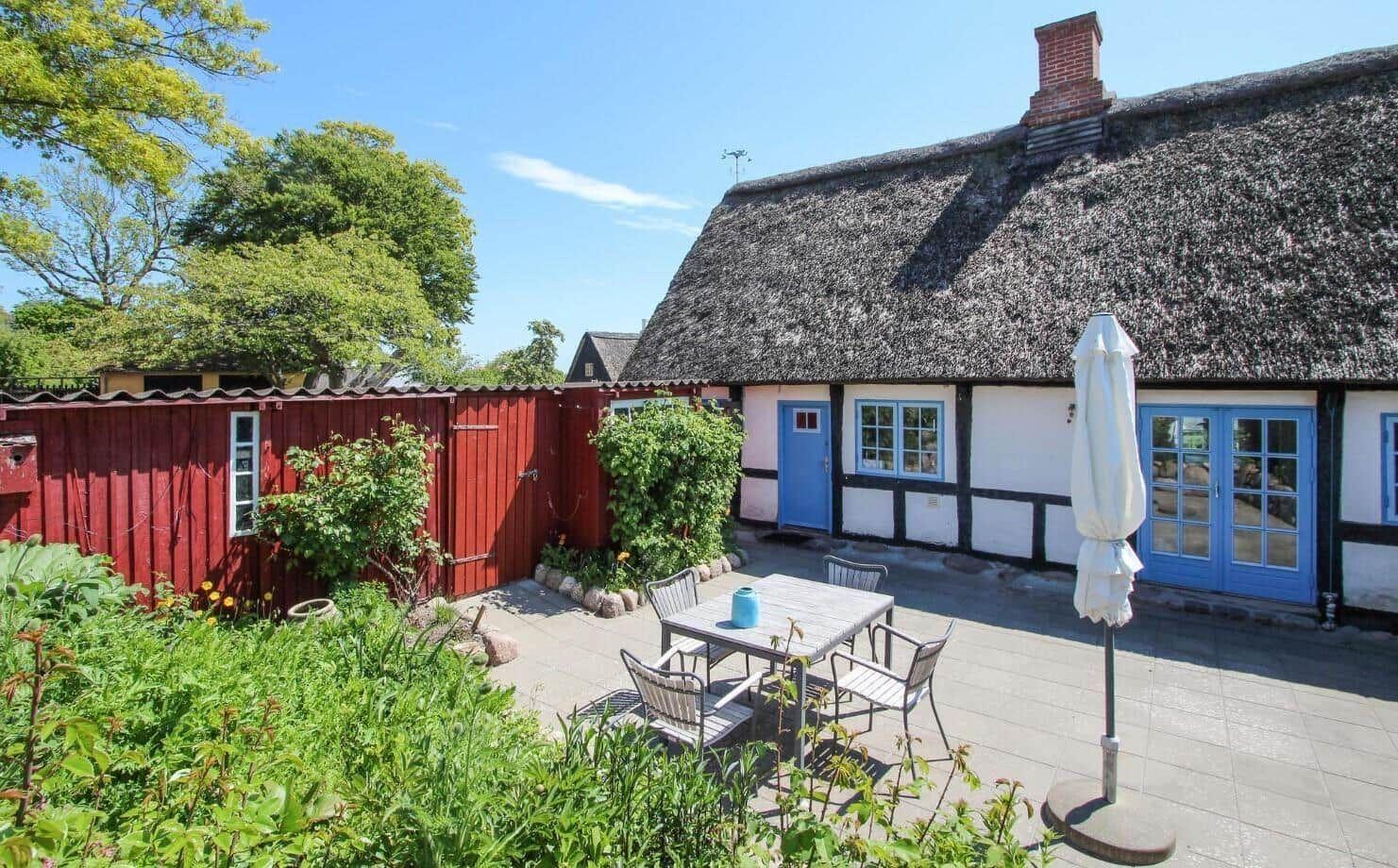 Hinterhof mit Tisch, Stühlen und geschlossenem Sonnenschirm. Umgeben von Grün und Steinmauern.