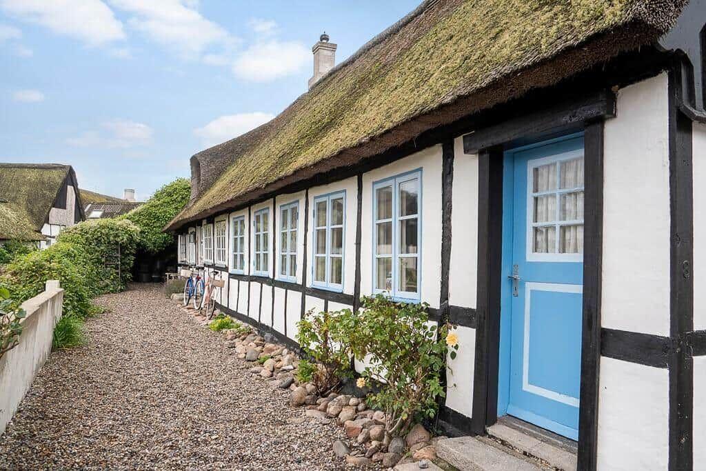 Seeufer mit Haus, Bäumen und Personen. Vordergrund: rosa Blumen.