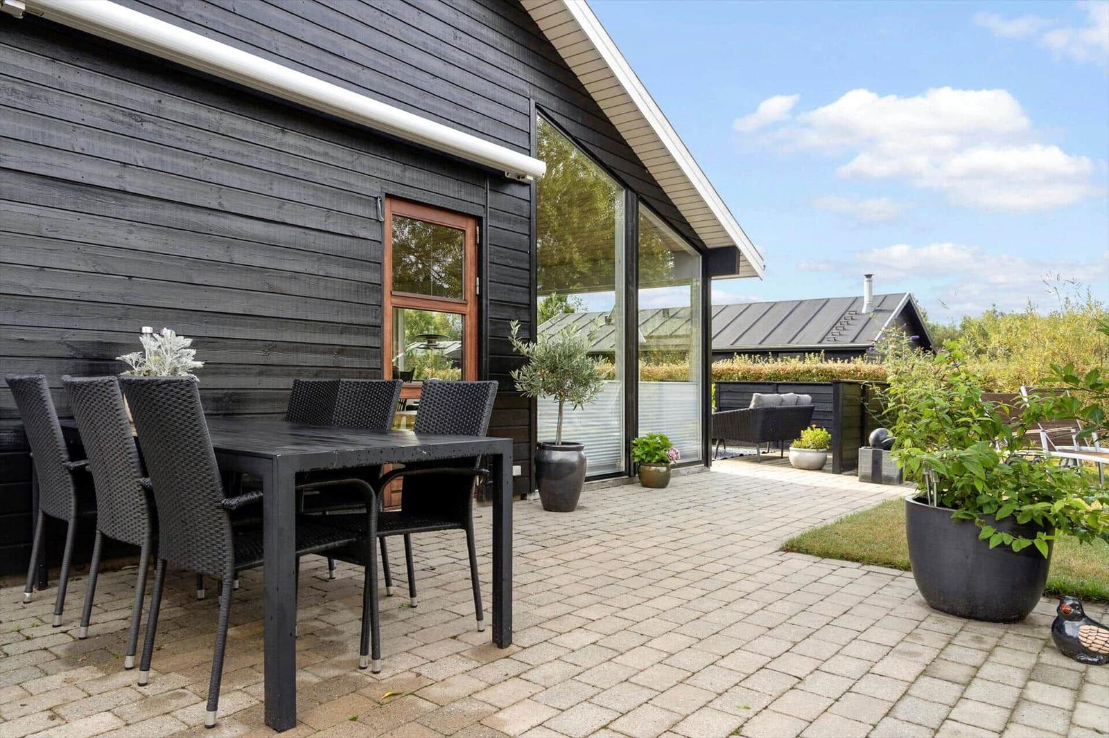 Terrace with seating, plants, and grill area. Background: wooden house and fence.