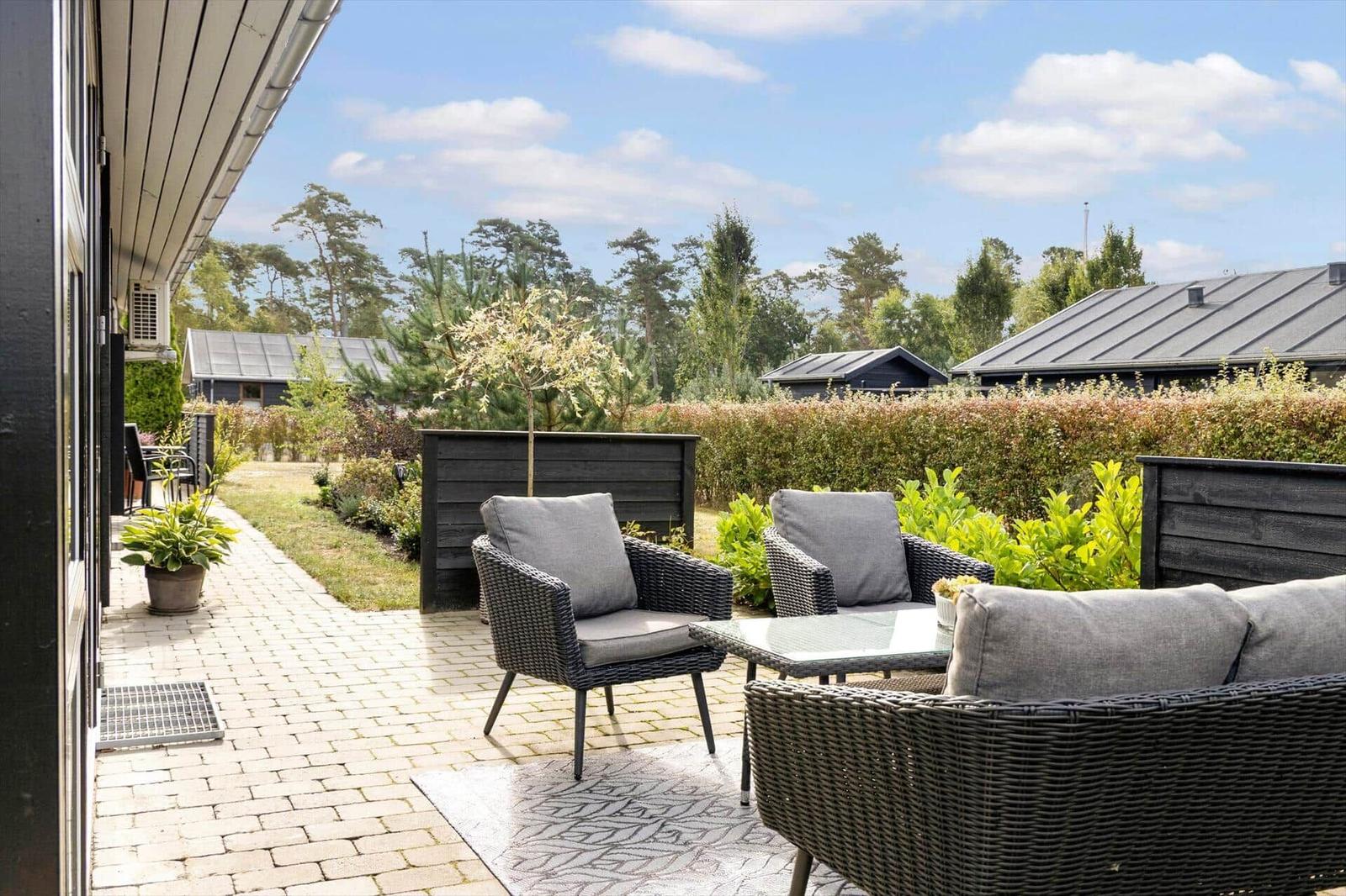 Terrace with table and chairs in front of a wooden house with garden and trees.