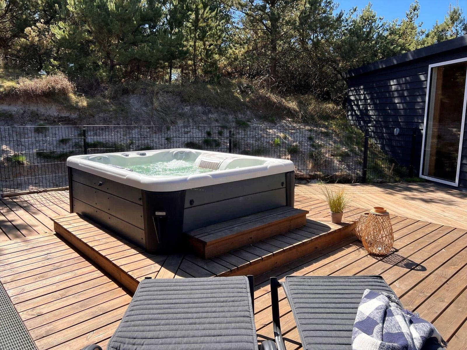 Terrasse mit Whirlpool, Sonnenliegen und Holzveranda. Hintergrund: Holzhaus mit Strohdach und Bäume.