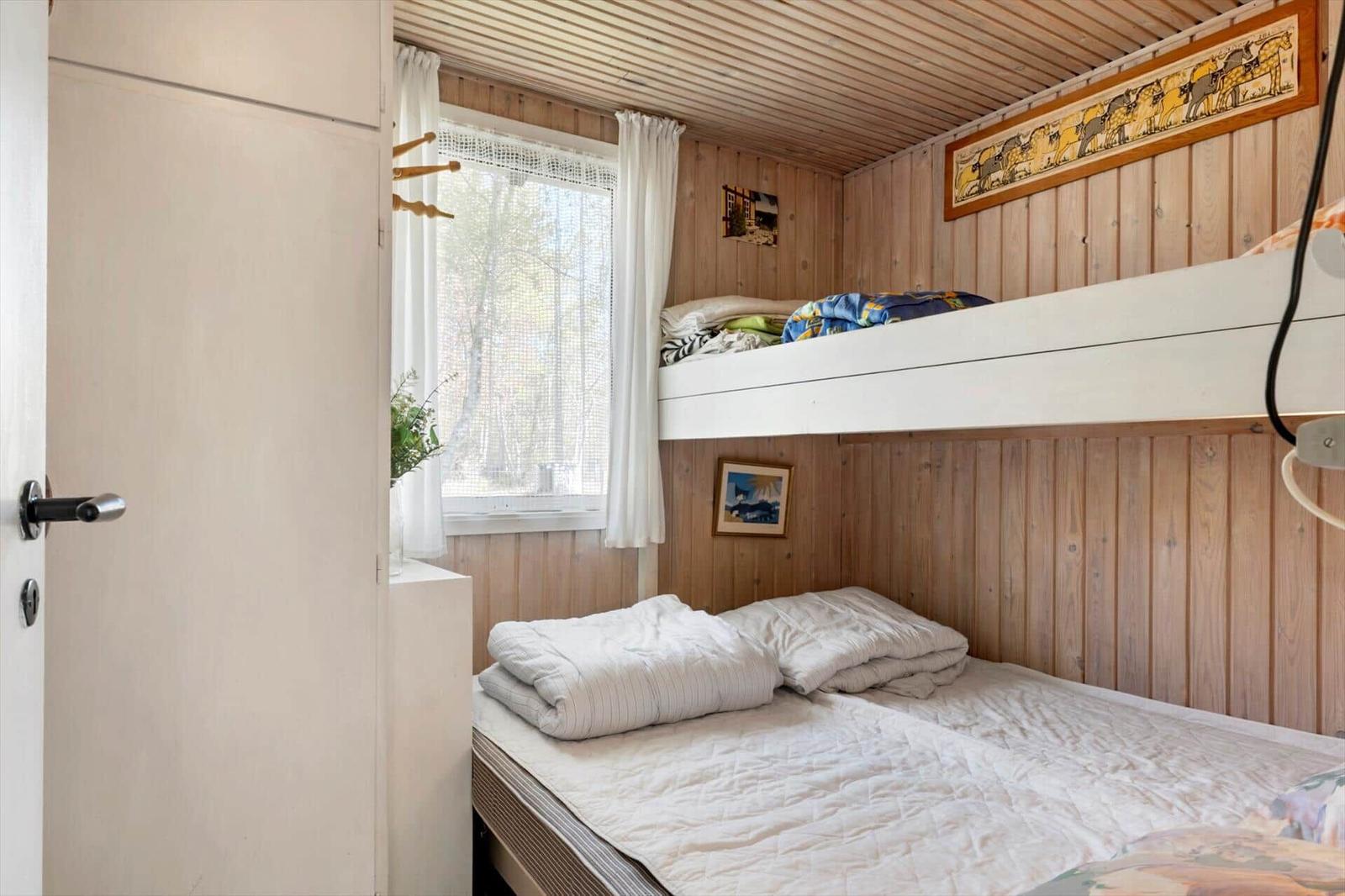 Schlafzimmer mit Doppelbett und obere Schlafcouch. Holzwände und Fenster mit weißen Vorhängen.