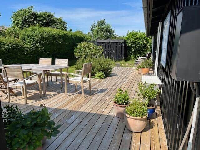 Schwarzes Holzhaus mit Terrasse, Garten und Bäumen. Tür offen, Sitzgelegenheit draußen.