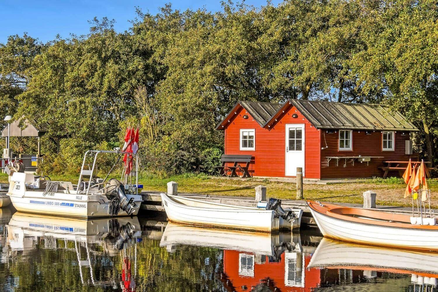 Rotes Bootshaus am Wasser mit drei Booten und Bäumen im Hintergrund.