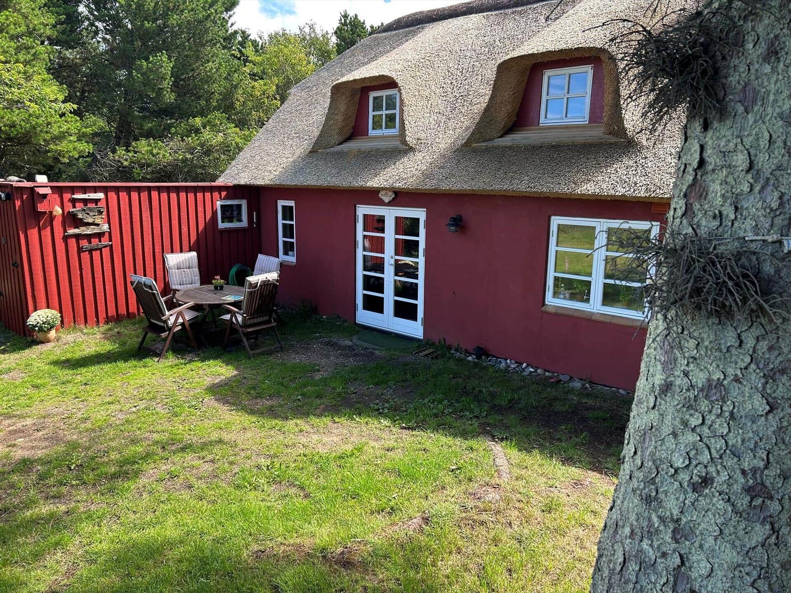 Rotes Haus mit Strohdach, Garten und Bäumen. Zwei Holzstühle vor der Tür.