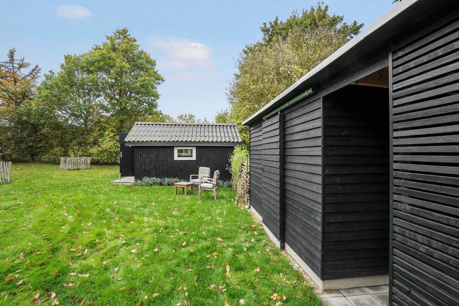 Schwarze Holzhütte mit Terrasse, Garten und Baum. Holzstapel und weitere Hütte im Hintergrund.