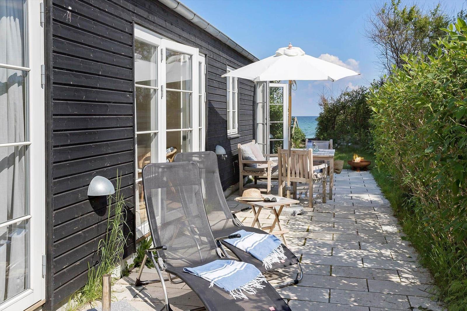 Schwarzes Holzhaus mit Garten und Meerblick. Tisch und zwei Stühle stehen draußen.