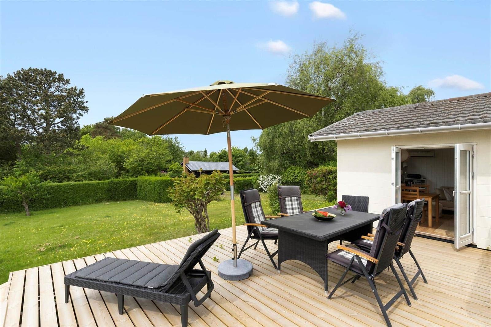 Deck with umbrella, lounge chairs, and table. Wooden steps lead to the deck. Background: house and garden.