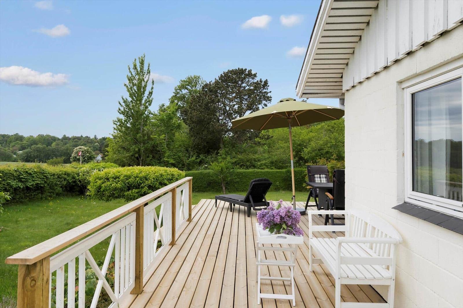 Terrace with table, chairs, and umbrella. Green background with hedges.