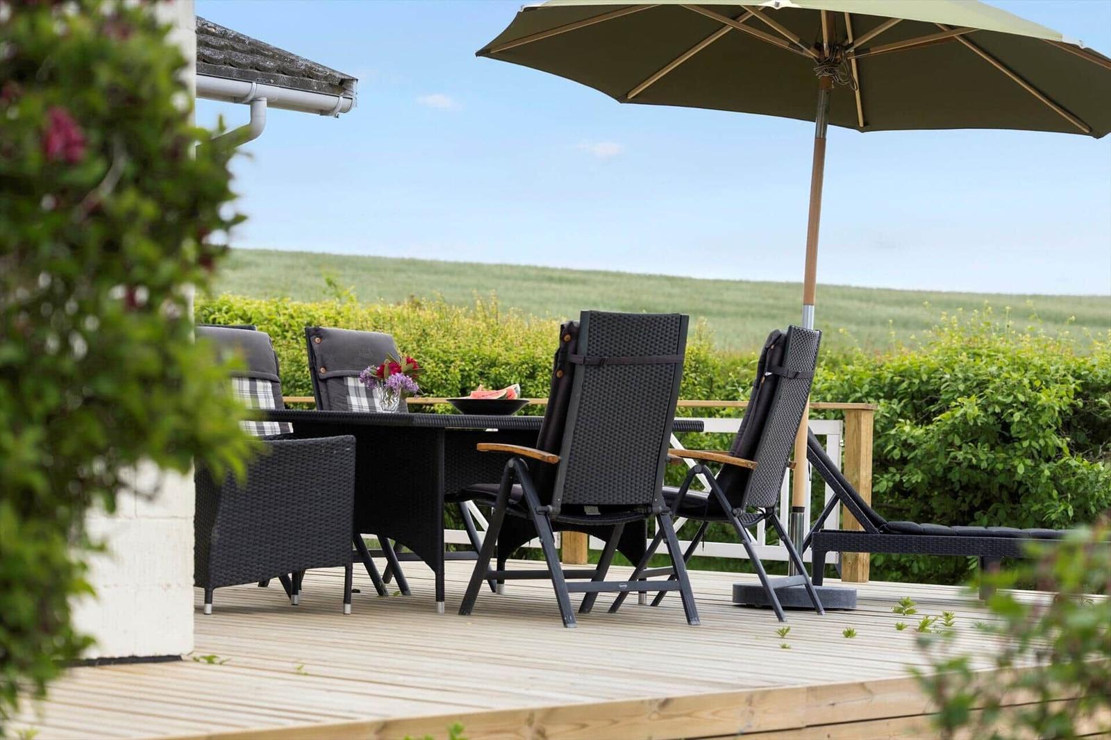 Deck with umbrella and lounge chairs. House in background. Table with watermelon and flowers.
