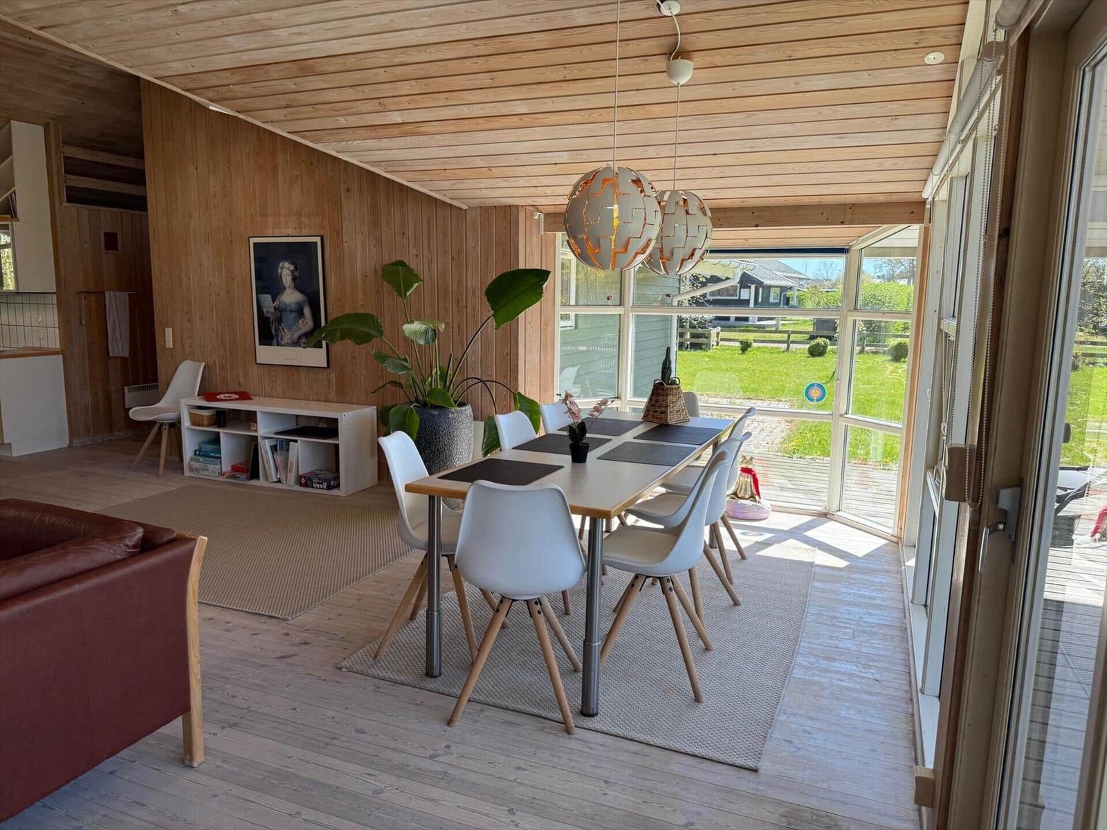 Essbereich mit Tisch, Stühlen und großen Fenstern. Holzwände und Decke. Pflanzen und Deckenleuchten.