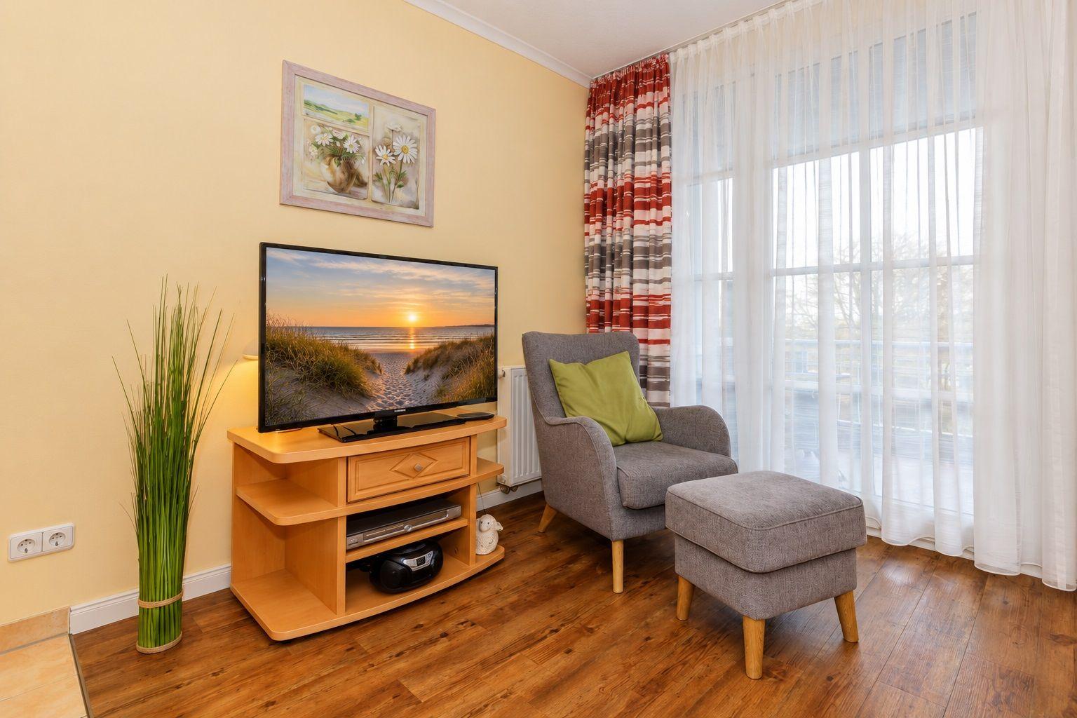 Ein Wohnzimmer mit TV, Sessel und Fußhocker. Fenster mit Vorhängen und Wandbild.