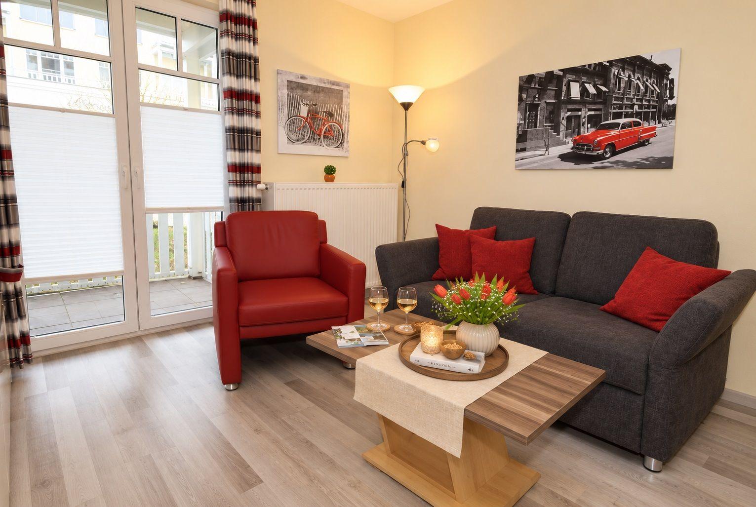 Wohnzimmer mit grauem Sofa, rotem Sessel und Tisch mit Wein. Fenster mit Jalousien und Wandbildern.