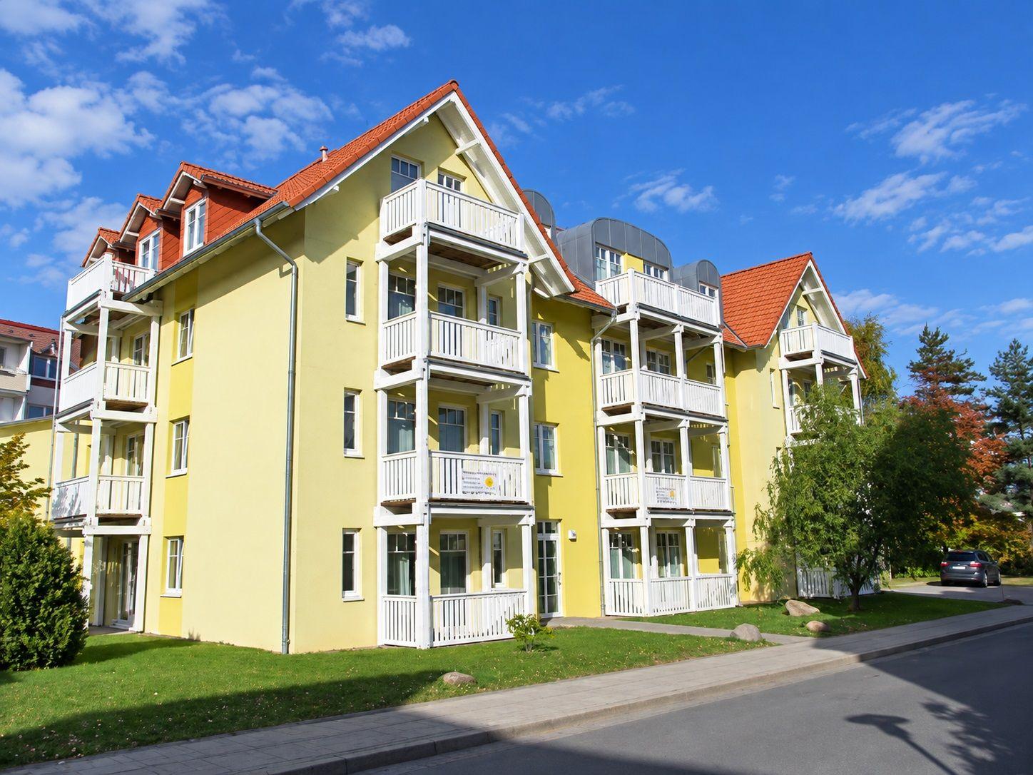 Gelbes Mehrfamilienhaus mit weißen Balkonen und rotem Dach unter blauem Himmel.