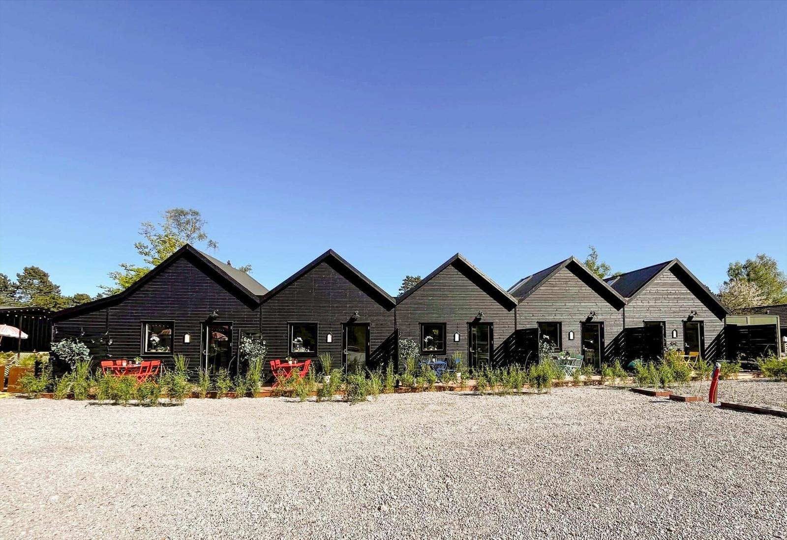 Row of dark wooden cabins with garden tables and clear sky.