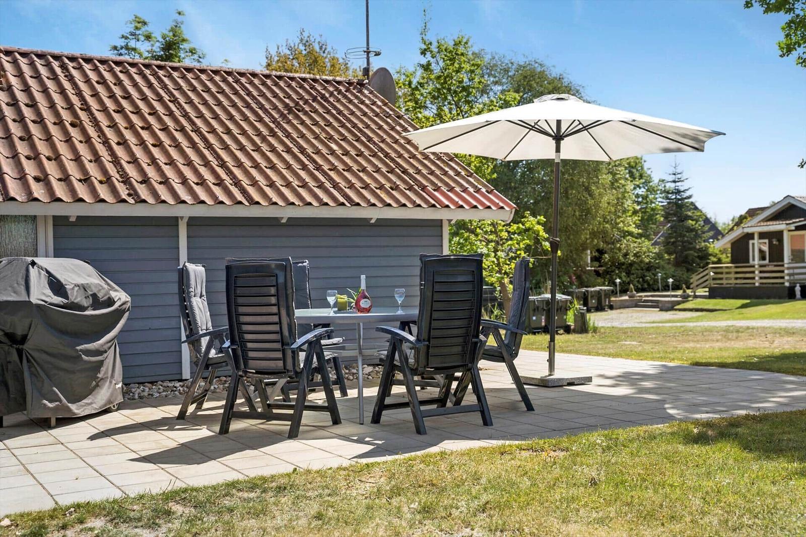 Terrasse mit Liege, Tisch und Stühlen unter Sonnenschirm. Grünfläche und Zaun im Hintergrund.