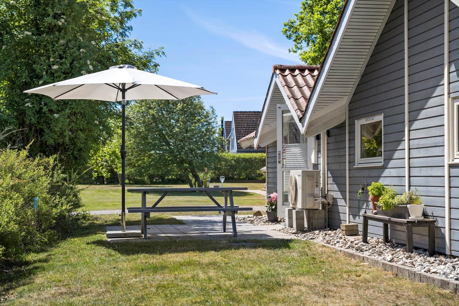 Ein graues Ferienhaus mit rotem Dach, Terrasse und Garten. Umgeben von Bäumen und einem Spielplatz.