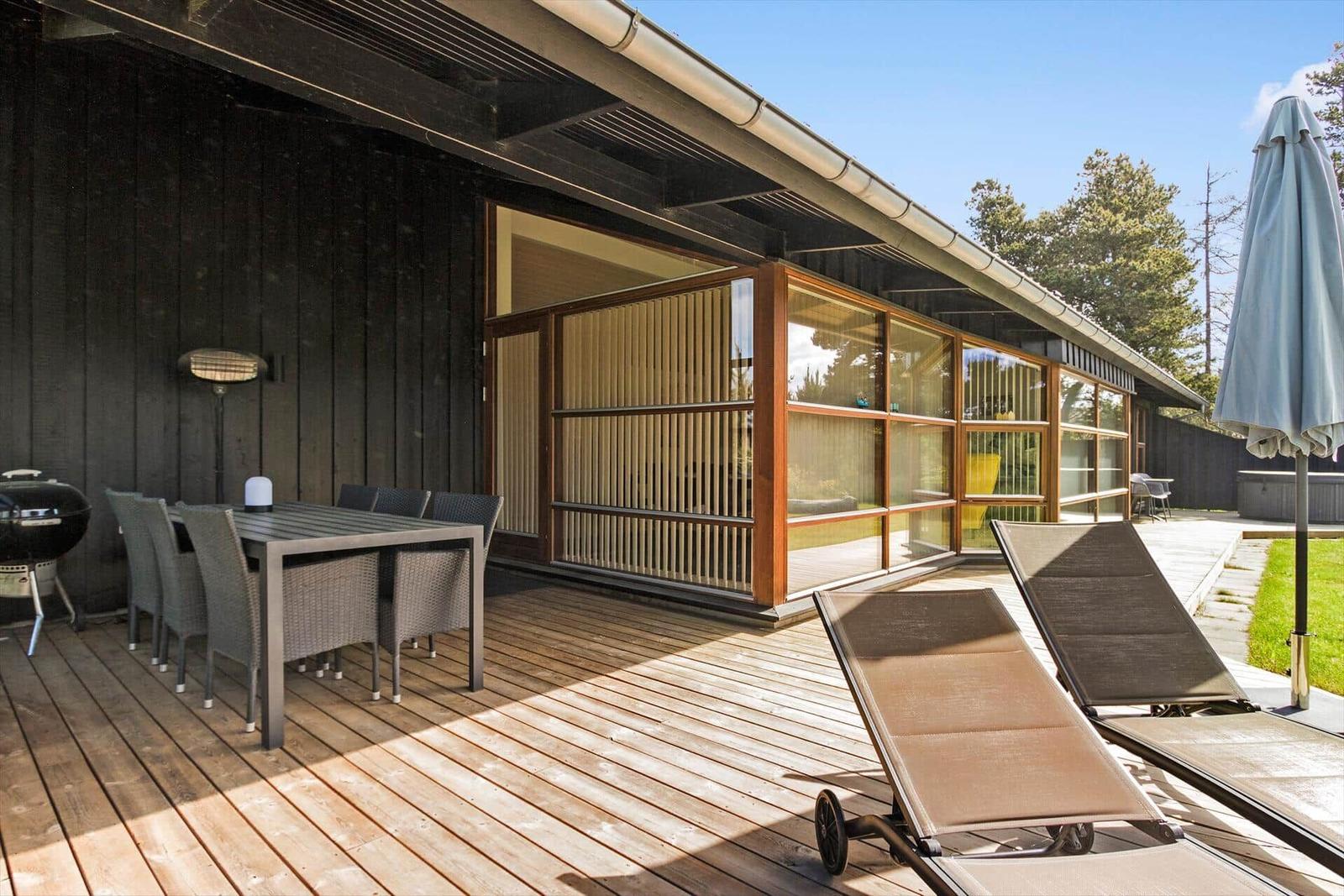 Terrasse mit Tisch, Stühlen, Liegestühlen und Grill. Holzwand und Glasfront sichtbar.