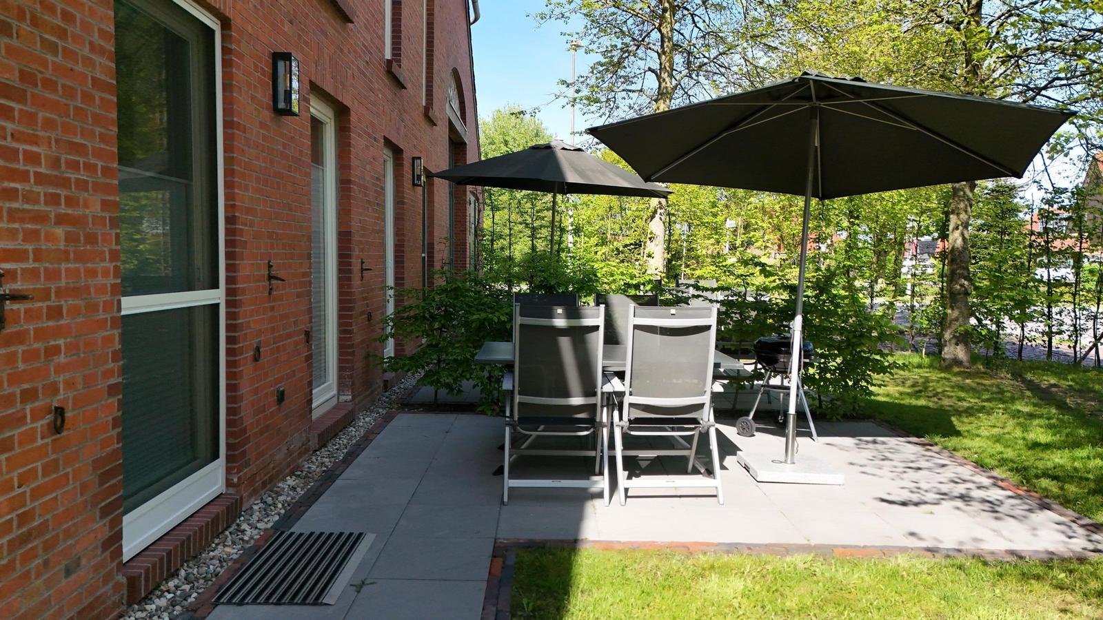 Terrasse mit Tisch, Stühlen, Sonnenschirm und Grill. Grünfläche und Backsteinhaus im Hintergrund.