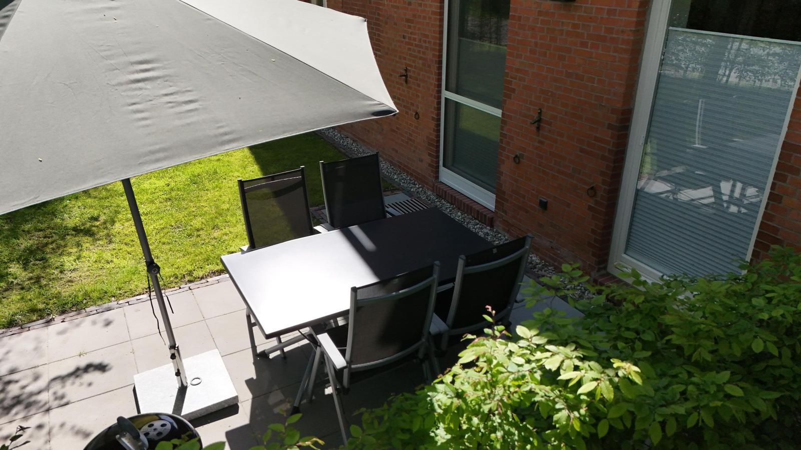 Terrasse mit Grill, Tisch, Stühlen und Sonnenschirm. Roter Backsteinbau und grüne Umgebung.