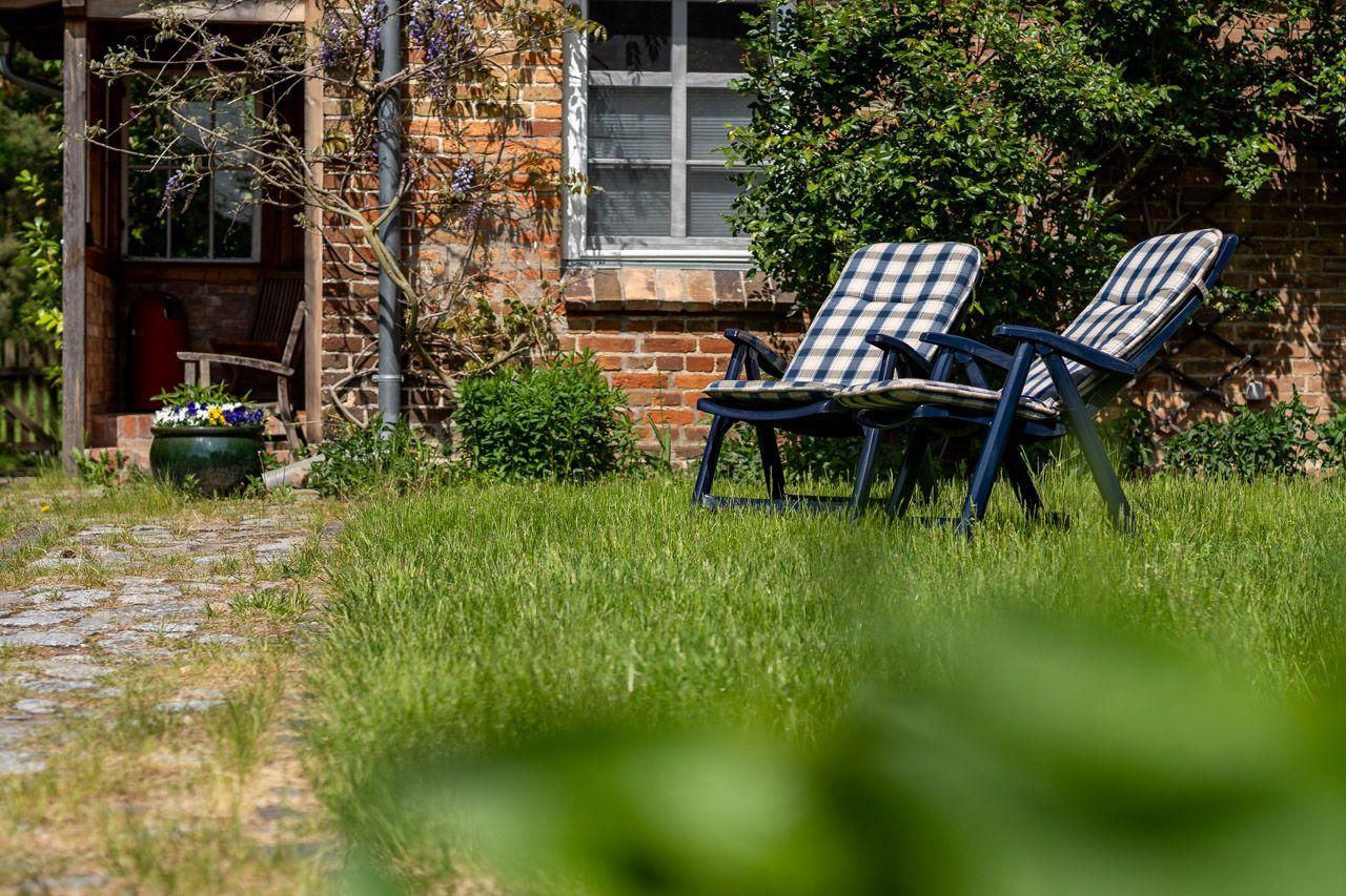 Grüner Garten mit Tisch, Stühlen und Sonnenschirm. Steinweg und Bäume im Hintergrund.