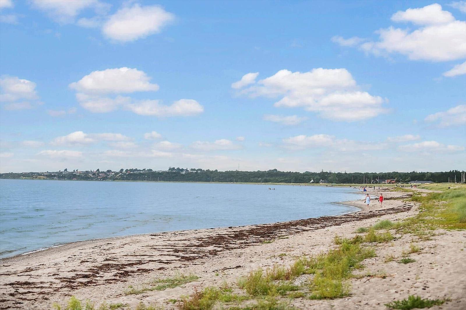 Beach with sand and grass, people walking along the water under blue sky with white clouds.