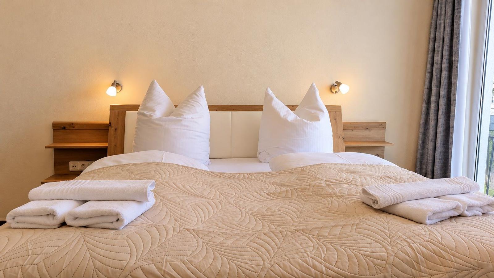 Doppelbett mit weißer Bettwäsche und gefalteten Handtüchern. Holzgestell und Wandlampen.