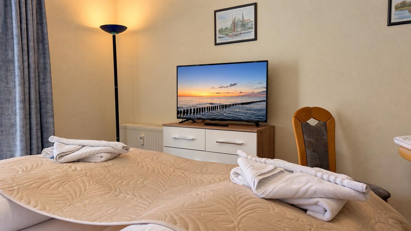 Schlafzimmer mit Bett, Fernseher und Stuhl. Auf dem Bett liegen Handtücher.