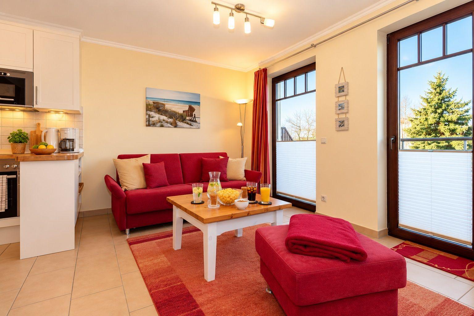Wohnbereich mit rotem Sofa, Holztisch und Fenster. Küche mit Arbeitsplatte und Geräten.