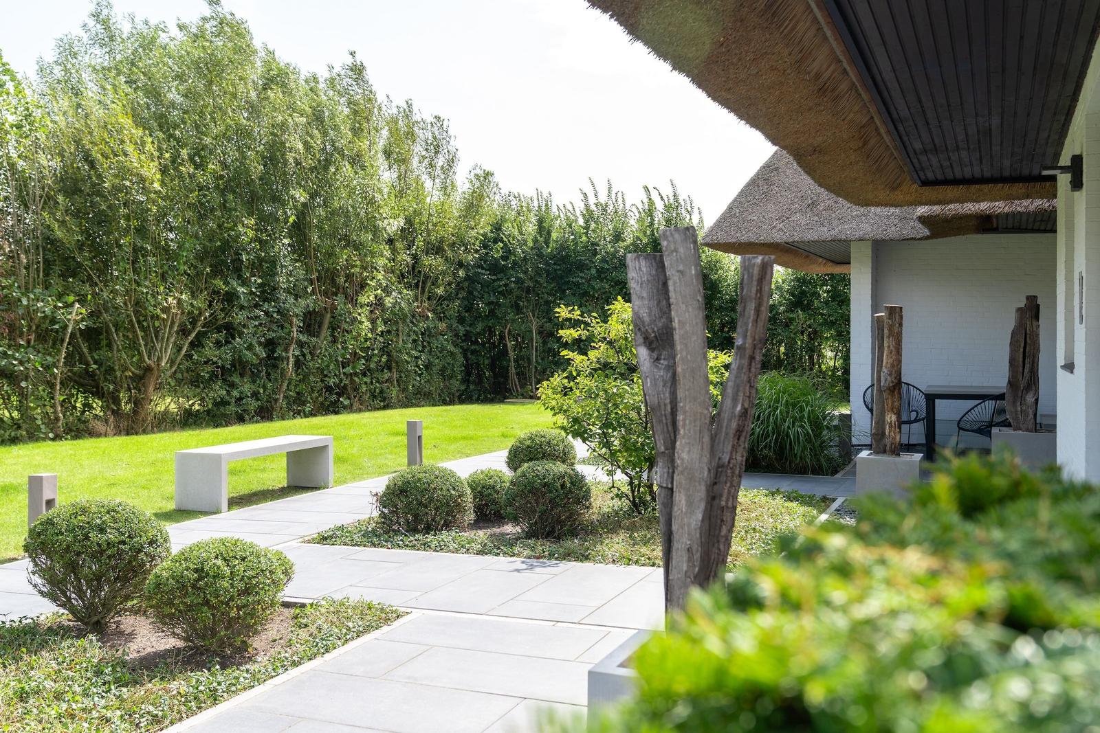 Garten mit Steinallee, grüner Rasen und moderner Bank unter strohgedecktem Vordach.