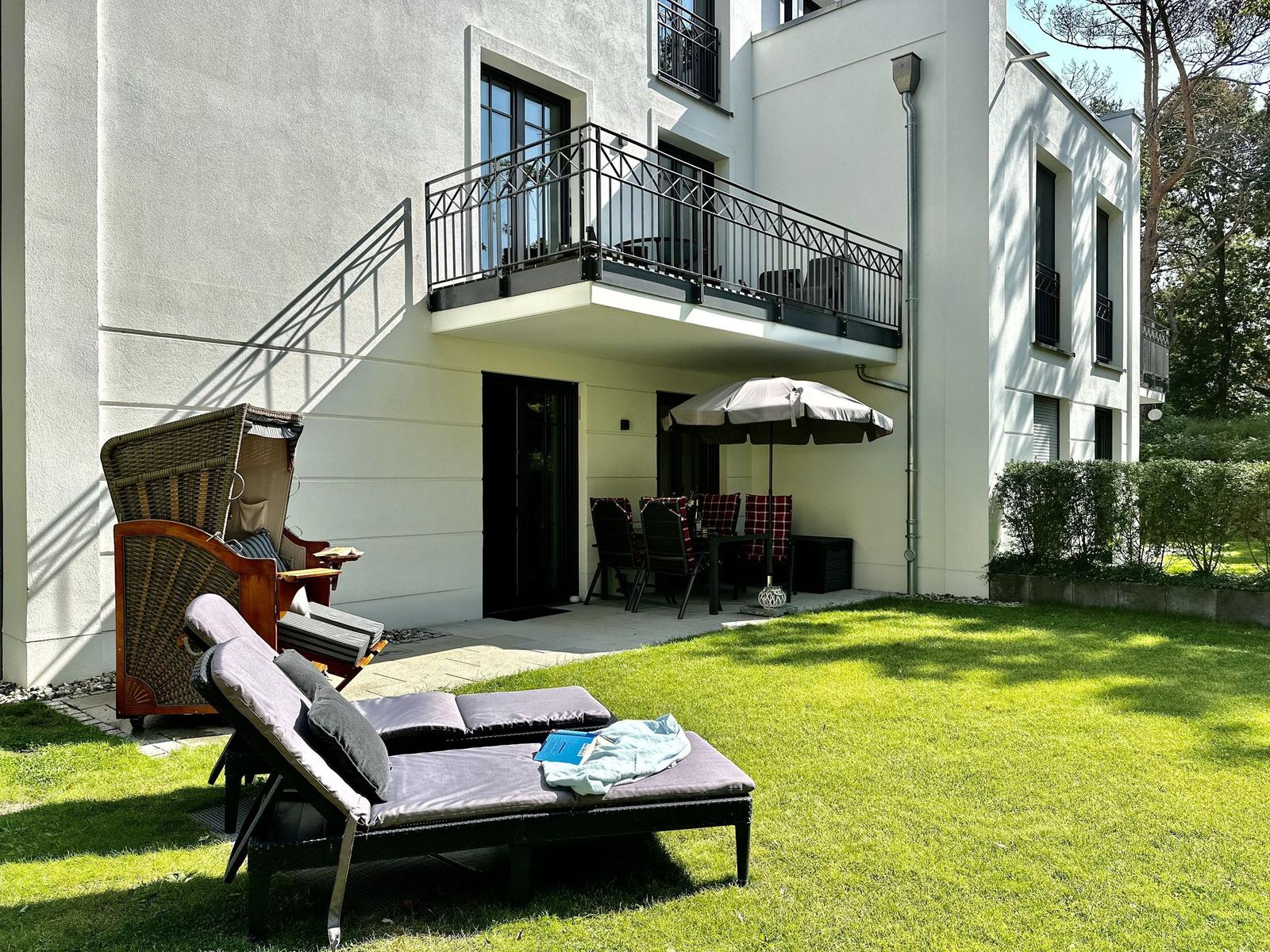 Terrasse mit Tisch, Stühlen und Sonnenschirm. Grünfläche und Gebäude im Hintergrund.
