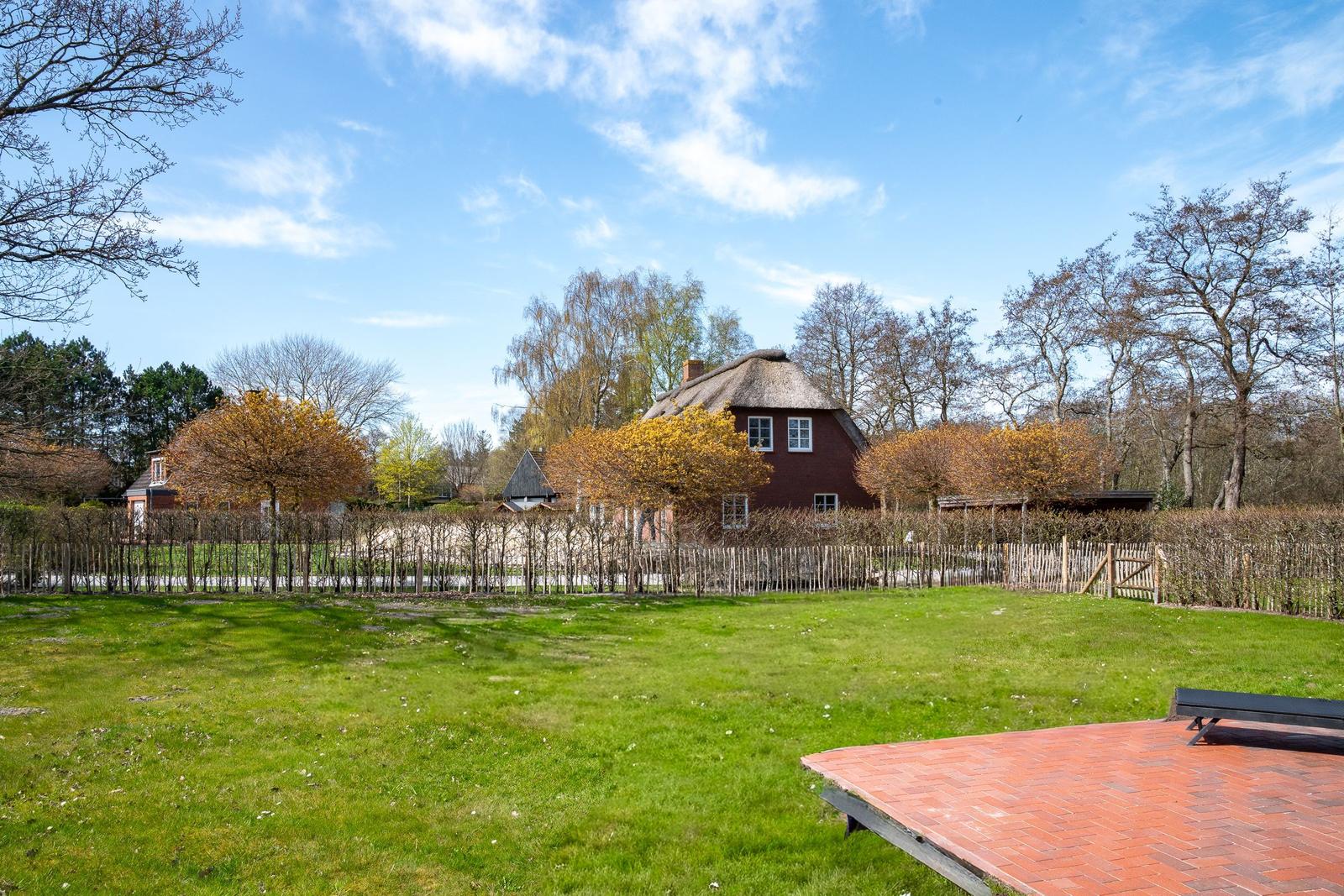 Roter Fachwerkhof mit Strohdach, umgeben von Grünflächen und Bäumen unter blauem Himmel.