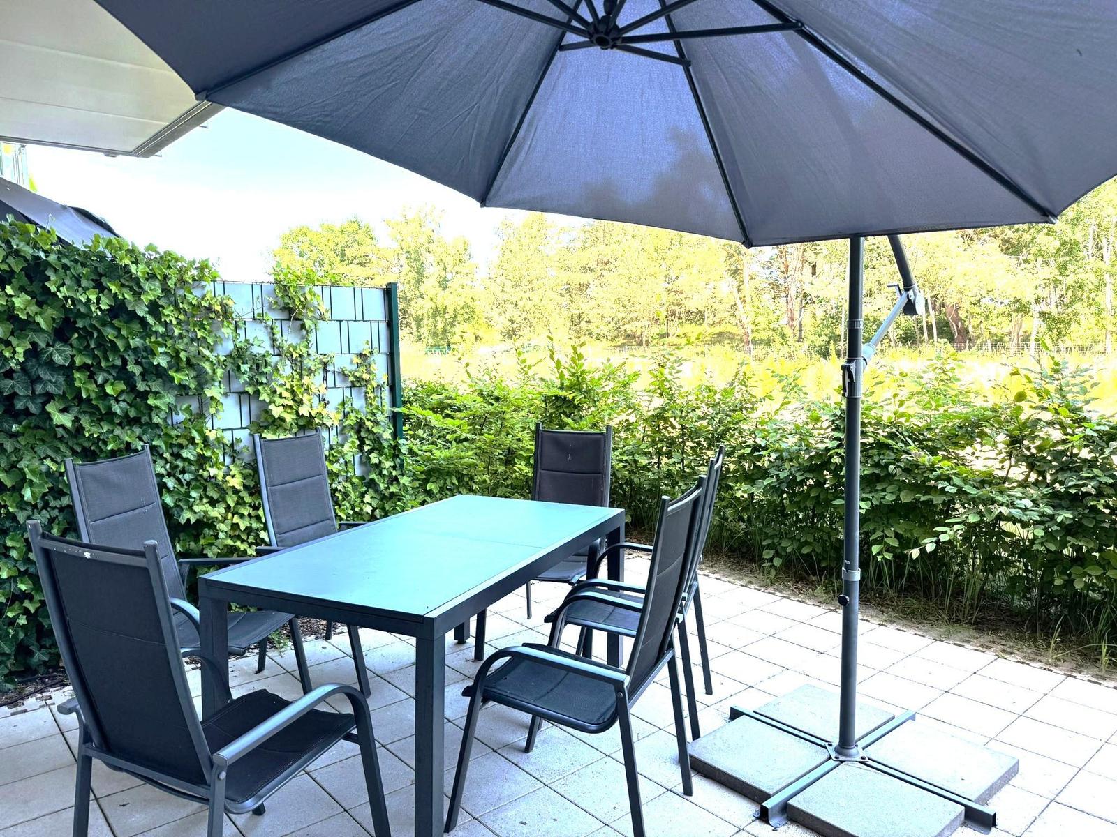 Terrasse mit Tisch, vier Stühlen und Sonnenschirm. Umgeben von grüner Hecke und Bäumen.