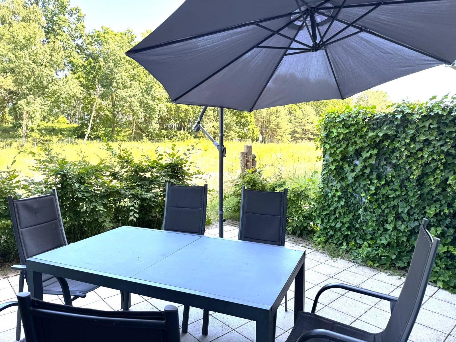 Terrasse mit Tisch, vier Stühlen und Sonnenschutz. Hintergrund: Grünflächen und Beerenhecke.