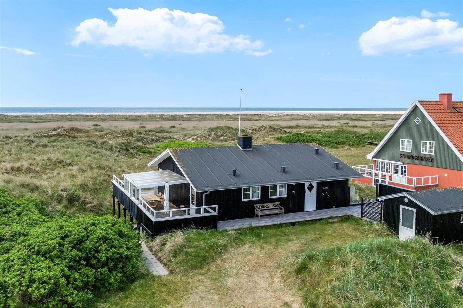 Schwarze Strandhütte mit Terrasse, umgeben von Dünen und Blick aufs Meer.