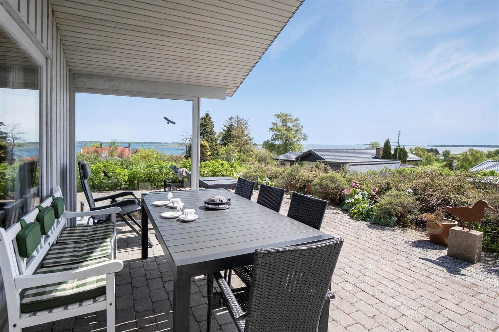 Terrasse mit Tisch, Stühlen und Liege. Blick auf Wasser und Grün. Helle, klare Wetterlage.