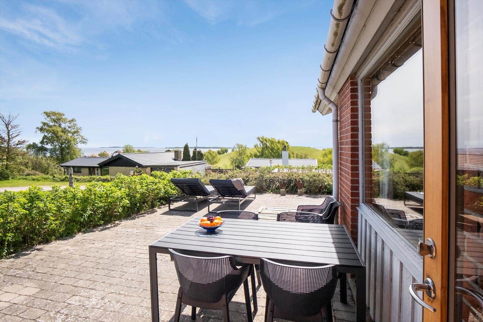 Terrasse mit Tisch, Stühlen und Liegestuhl. Blick auf Wasser und Grünflächen.