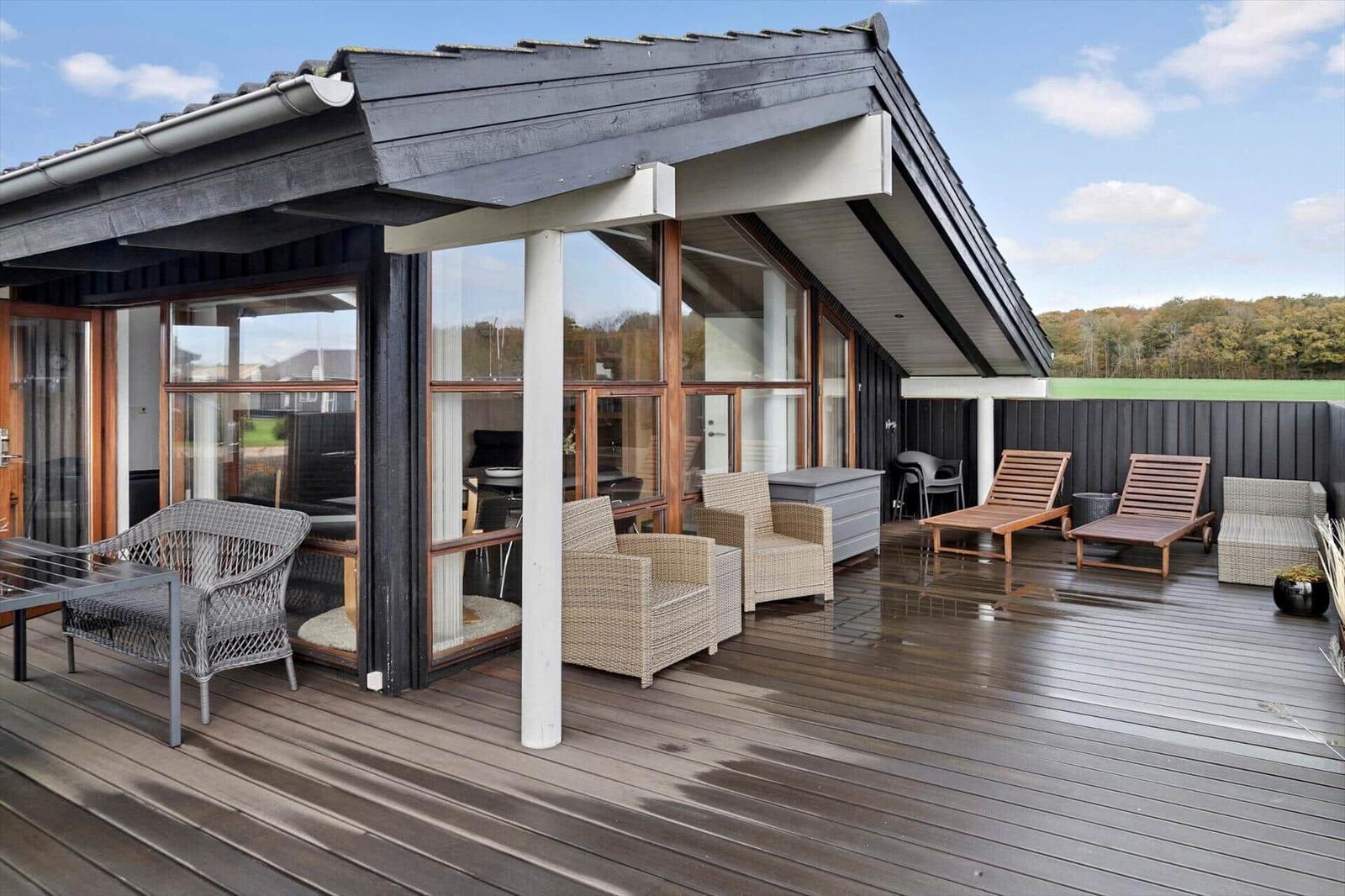Haus mit großem Holzterrasse, Grill und Gartenmöbeln. Blick auf grünes Feld.